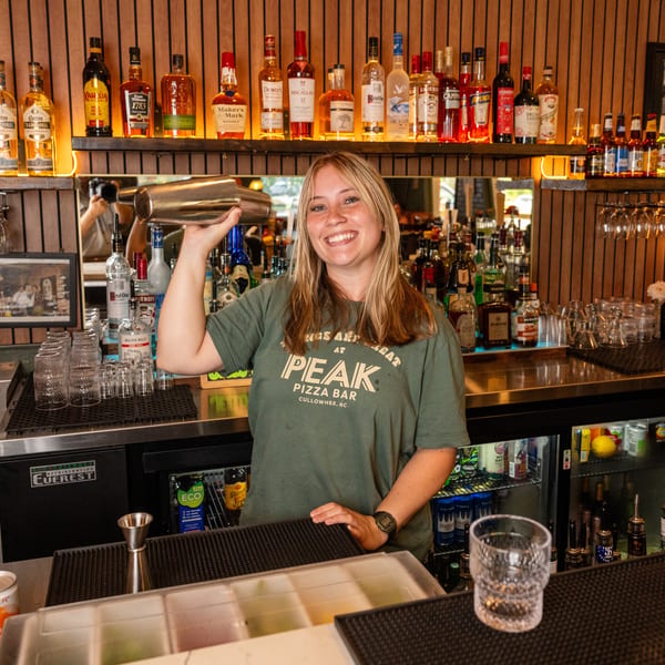Smiling bartender in a Peak Pizza Bar shirt shaking a cocktail behind a well-stocked bar.