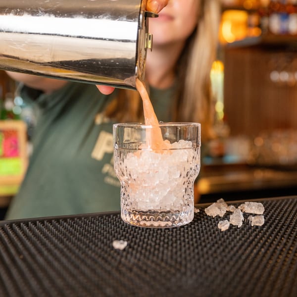 Bartender pouring a pink cocktail from a shaker into a glass filled with crushed ice.