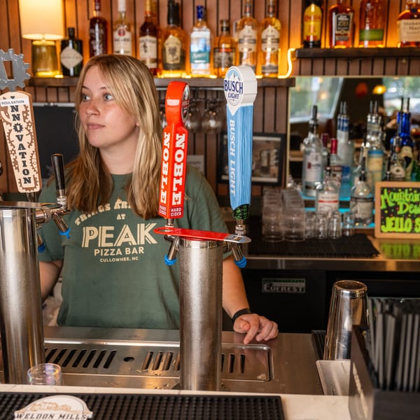 A bartender at Peak Pizza Bar stands behind a bar with beer taps and liquor bottles.