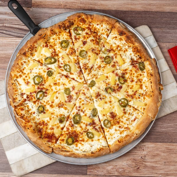 A sliced pizza with white sauce, pineapple, and jalapeños on a metal pan with a server.