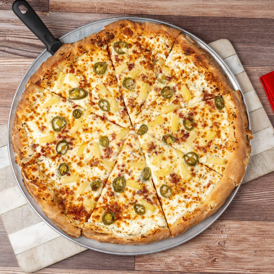 A sliced pizza with white sauce, pineapple, and jalapeños on a metal pan with a server.