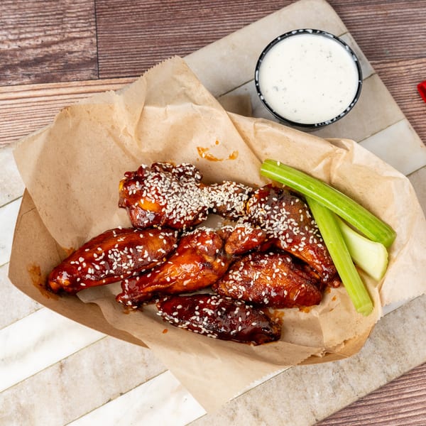 Sweet and spicy chicken wings with sesame seeds, celery sticks, and ranch dressing on a wooden table.