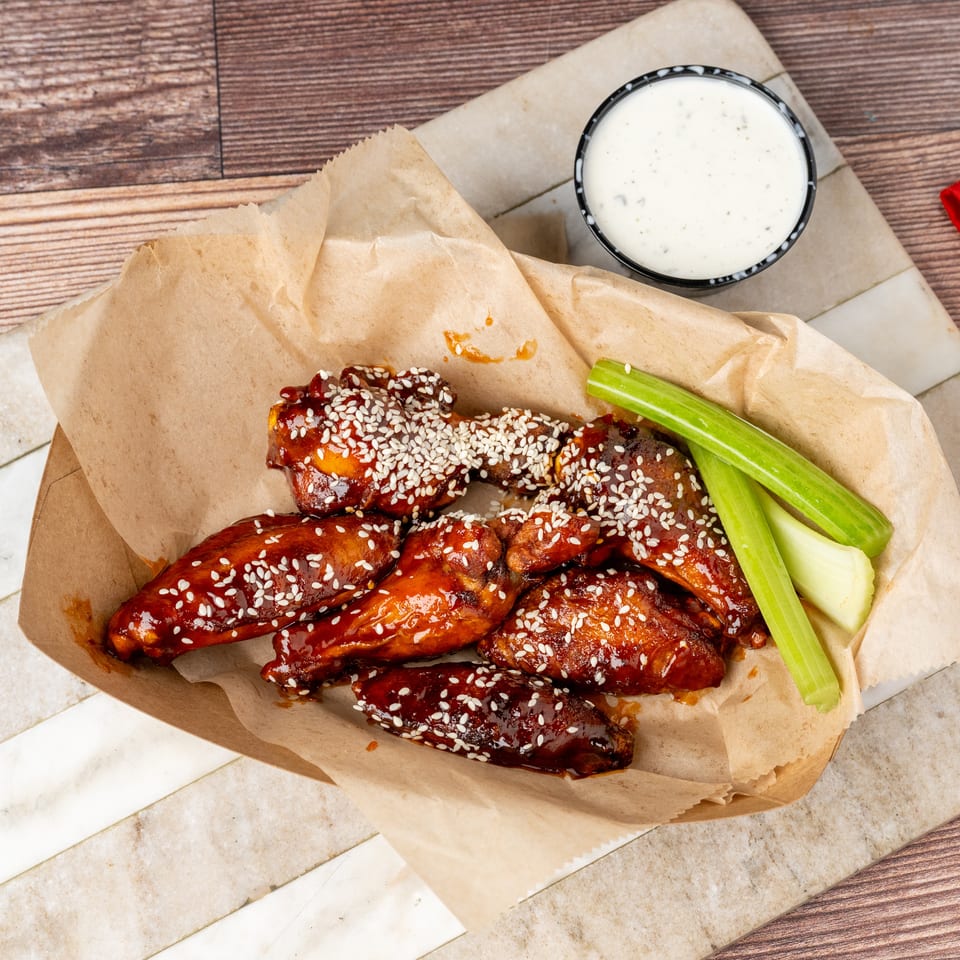 Sweet and spicy chicken wings with sesame seeds, celery sticks, and ranch dressing on a wooden table.