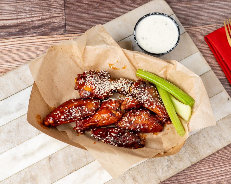 Sweet and spicy chicken wings with sesame seeds, celery sticks, and ranch dressing on a wooden table.