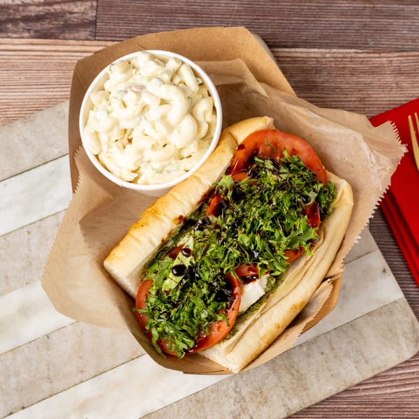 A sub sandwich with lettuce, tomato, and balsamic glaze, next to a cup of macaroni salad.
