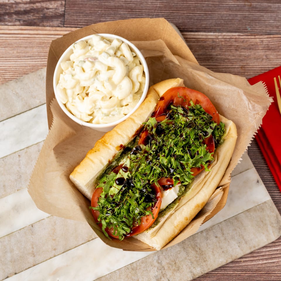 A sub sandwich with lettuce, tomato, and balsamic glaze, next to a cup of macaroni salad.