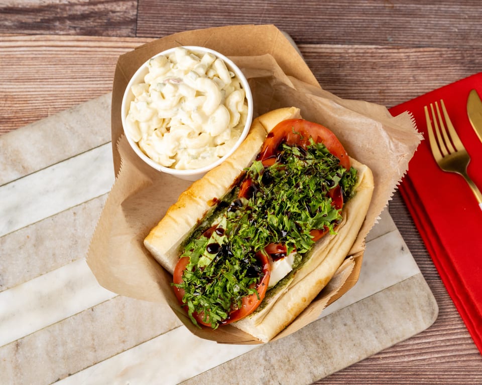 A sub sandwich with lettuce, tomato, and balsamic glaze, next to a cup of macaroni salad.