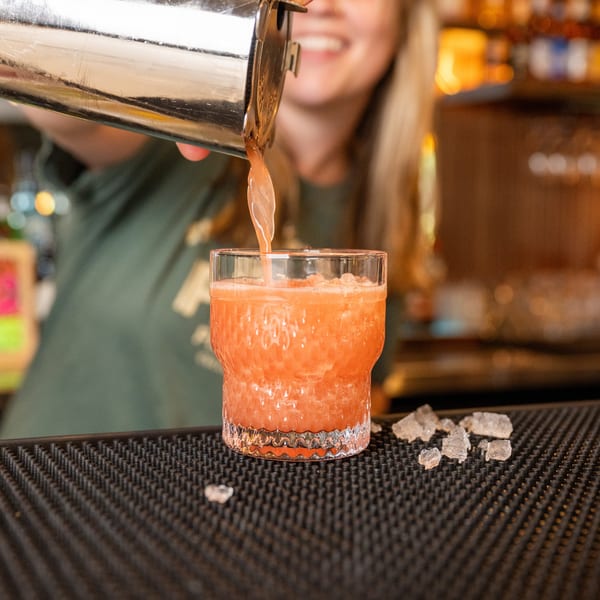 Bartender pouring a pink cocktail from a shaker into a glass with ice on a bar mat.