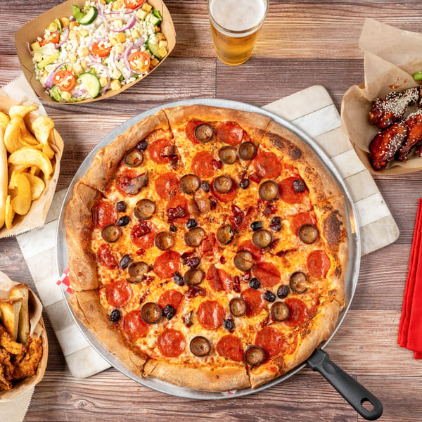 A pepperoni and sausage pizza with various side dishes, drinks, and cutlery on a wooden table.