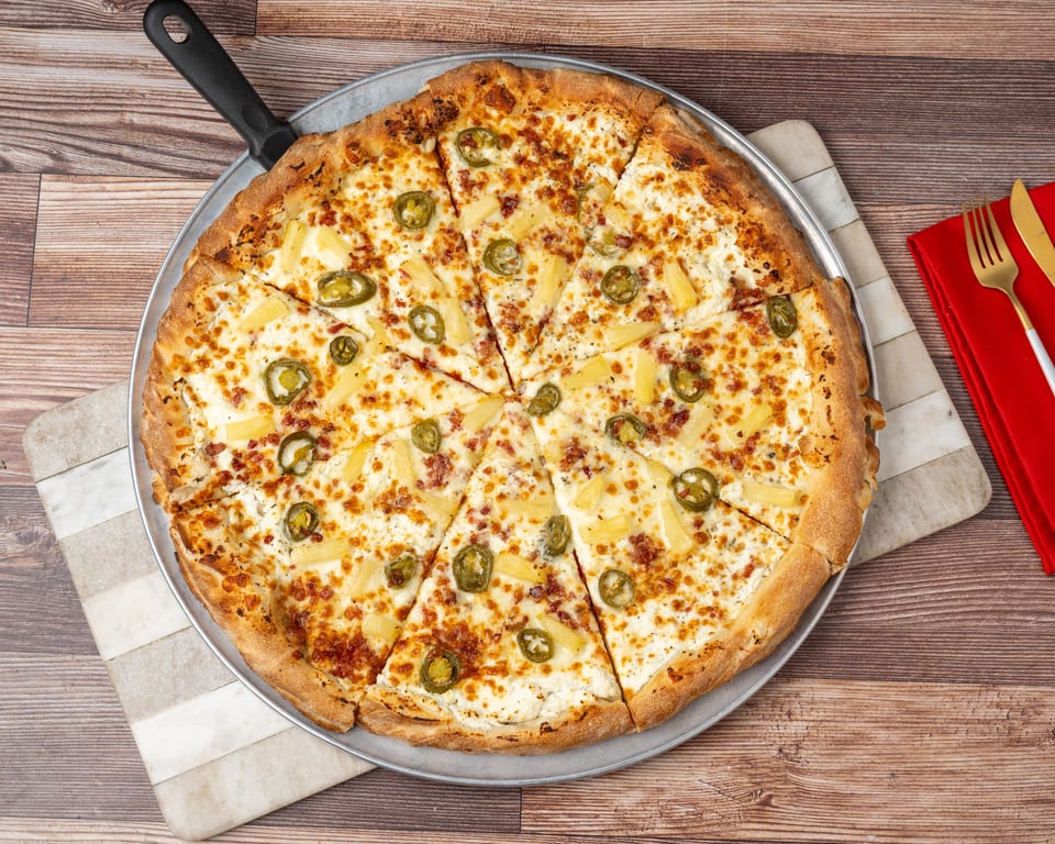 A whole pizza with white sauce, pineapple, jalapeños, and bacon bits, sliced on a metal pan.