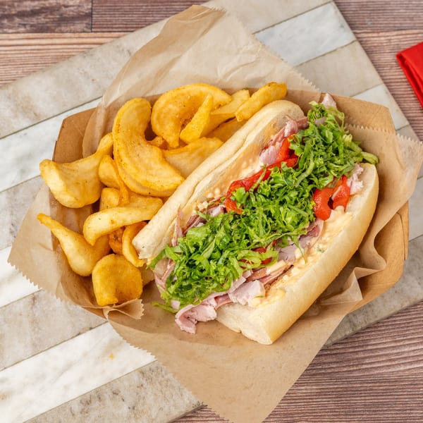 A sub sandwich with lettuce, red peppers, and meat, served with potato chips in a paper tray.