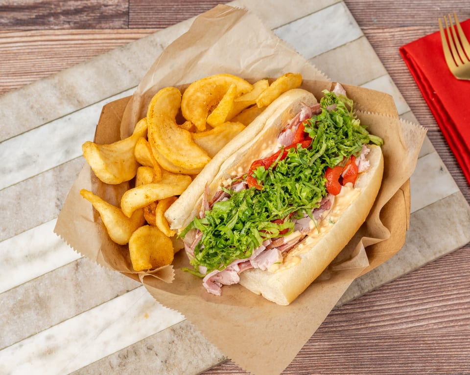 A sub sandwich with lettuce, red peppers, and meat, served with potato chips in a paper tray.