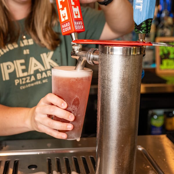 A bartender in a Peak Pizza Bar shirt pours a hard cider from a tap into a frosty glass.