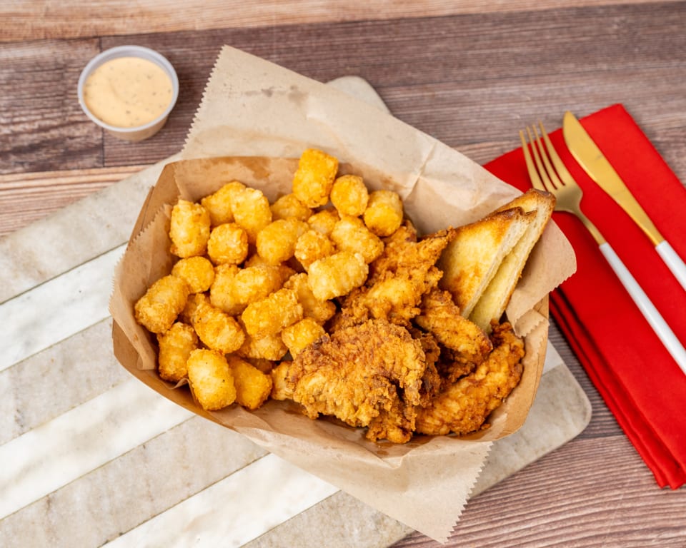 Fried chicken tenders, tater tots, and Texas toast in a paper-lined basket with dipping sauce.