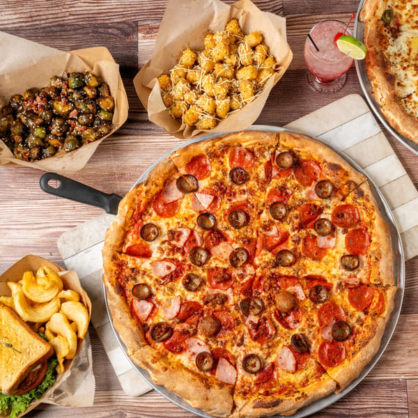A wooden table laden with two pizzas, a sandwich with fries, brussels sprouts, and tater tots.