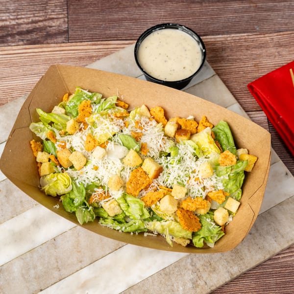 Caesar salad with croutons and shredded cheese in a brown paper container, with dressing on the side.