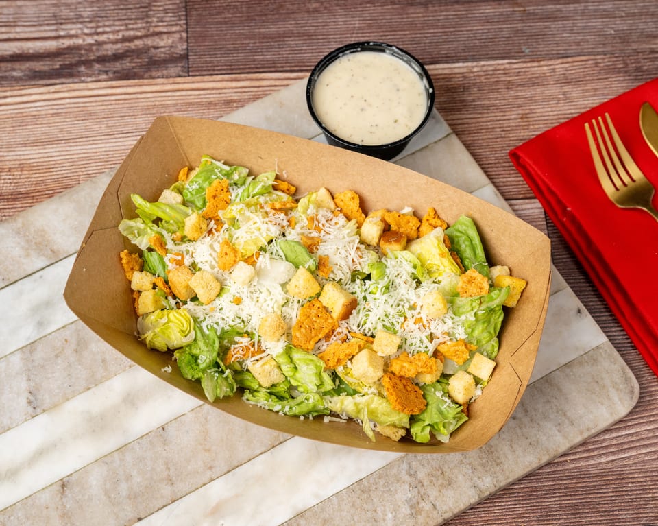 Caesar salad with croutons and shredded cheese in a brown paper container, with dressing on the side.