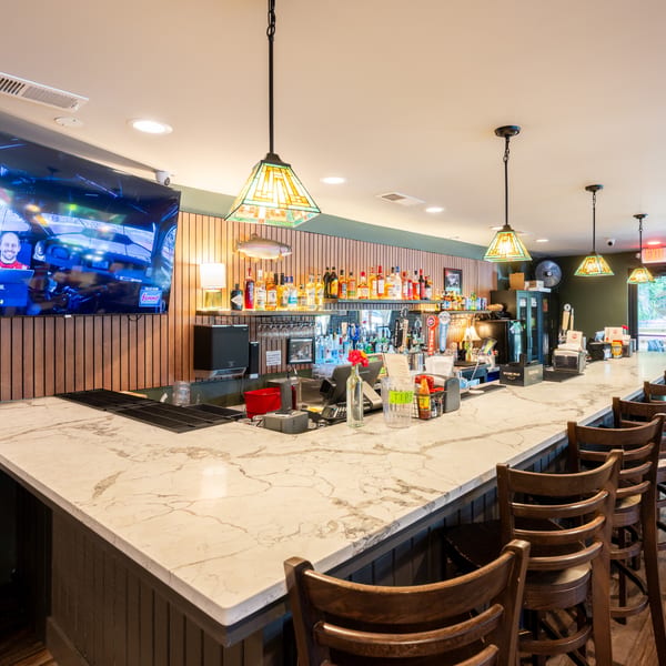 A well-lit bar area with a marble countertop, wooden bar stools, and a large TV screen.