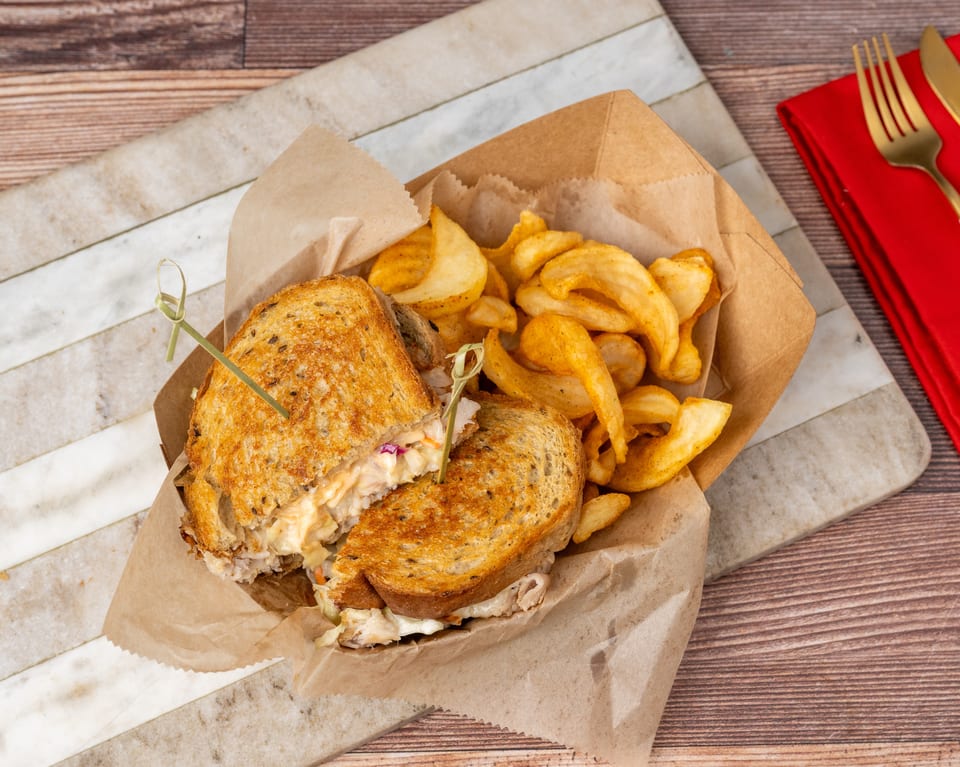 Grilled sandwich with coleslaw and fries in a paper tray, on a marble and wood surface.